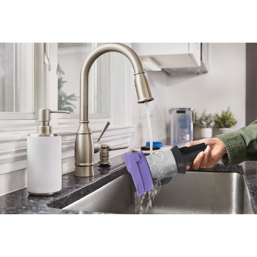 Photo of a BLACKDECKER BSV520BRP vacuum filter with purple cap being rinsed under a kitchen faucet in a stainless steel sink.