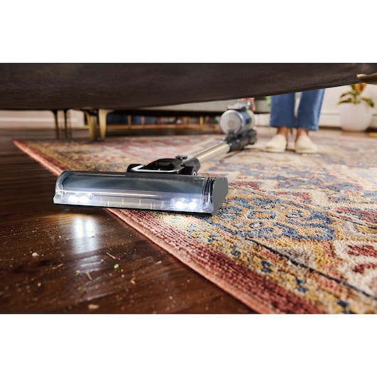 BLACKDECKER BSV525BLP cordless vacuum cleaning under furniture on a patterned rug and hardwood floor.