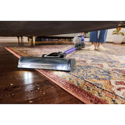 BLACKDECKER BSV525BRP cordless stick vacuum cleaning under furniture on a patterned rug and hardwood floor, close-up angle.