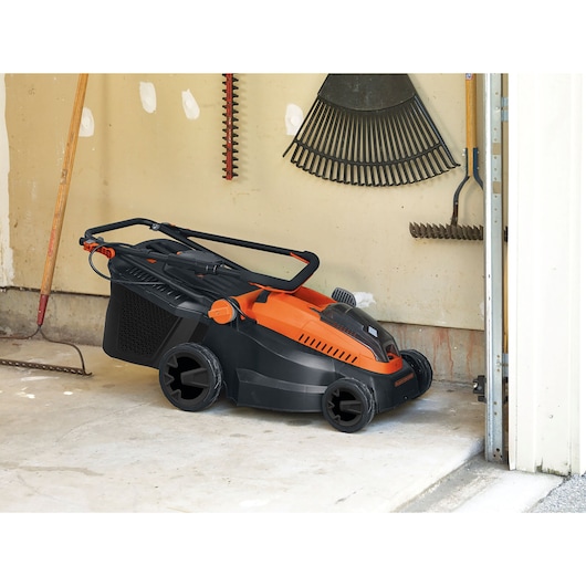 BLACK+DECKER CLM3825L2 lawn mower in black and orange, folded for storage in a garage next to garden tools.