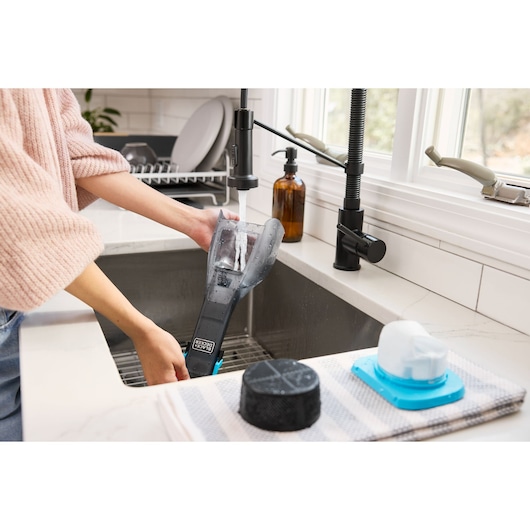Photo of a person rinsing BLACK+DECKER DVD320B vacuum parts at a kitchen sink with accessories on a striped towel.