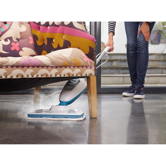BLACK+DECKER FSM13E1 steam mop cleaning dark floor under patterned armchair, handled by person in jeans.
