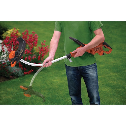 A person is holding a BLACK+DECKER electric grass trimmer in a garden, demonstrating its adjustable head feature.