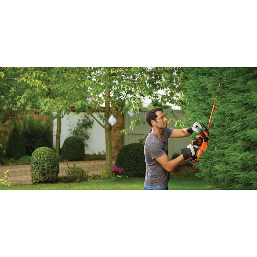 Person trimming tall hedge with BLACKDECKER cordless hedge trimmer GTC18452PC in a garden.