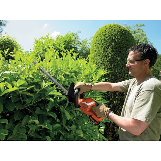 BLACK+DECKER hedge trimmer GTC1845L20 in use trimming green bushes outdoors, operator wearing gloves.