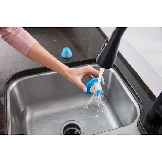 BLACKDECKER HLVC320B11 scrub brush part being rinsed under faucet in stainless steel kitchen sink.