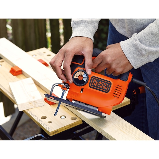 BLACK+DECKER KS801SE jigsaw in use, orange and black, cutting wood on a workbench.