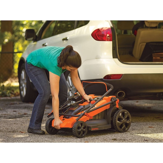 Person loading a folded BLACK+DECKER LM2000 lawn mower near white SUV in driveway.