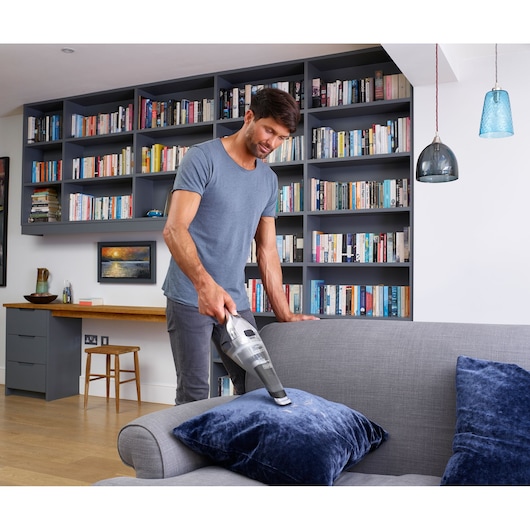 BLACKDECKER NVC220WC handheld vacuum cleaner being used on a grey sofa with blue cushions in a modern living room.