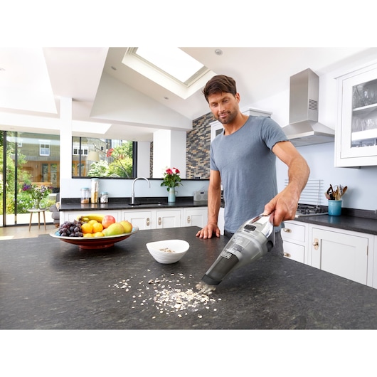 Photo of BLACK+DECKER NVC220WC handheld vacuum cleaning spilled oats on a kitchen countertop near a bowl and fruit plate.