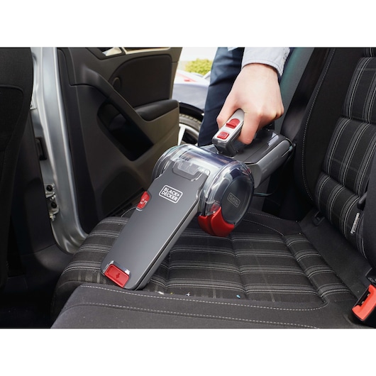 A BLACK+DECKER handheld vacuum cleaner is being used to clean the seat inside a car. The vacuum is grey with red accents and held by a person’s hand.