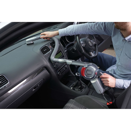A person is using a BLACK+DECKER handheld vacuum cleaner (model PV1200AV) with an extended hose to clean the dashboard and interior of a car.