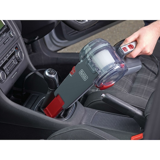 A BLACK+DECKER handheld vacuum cleaner (model PV1200AV) is being used to clean the interior of a car, specifically around the gear shift area.