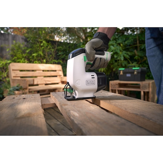 BLACKDECKER REVJ12C jigsaw cutting wood outdoors, gloved hand holding tool, outdoor furniture in background.