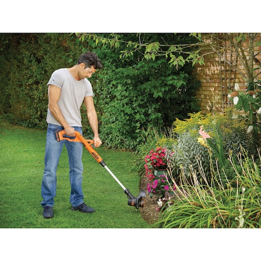 BLACK+DECKER ST182320 grass trimmer used in garden, orange and black cordless tool near flower bed.