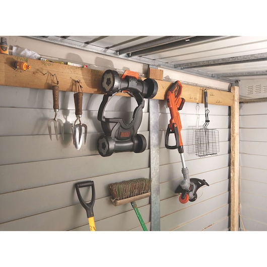 BLACKDECKER STC1820CM cordless grass trimmer and mower attachment hanging on shed wall with garden tools.