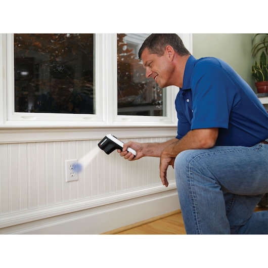 Photo of a person using a BLACKDECKER TLD100 thermal leak detector near a wall outlet in a home.