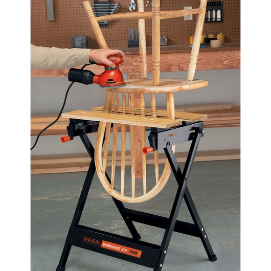 A wooden chair is being sanded using an electric sander, supported by a BLACK+DECKER Workmate 301 portable workbench in a workshop.