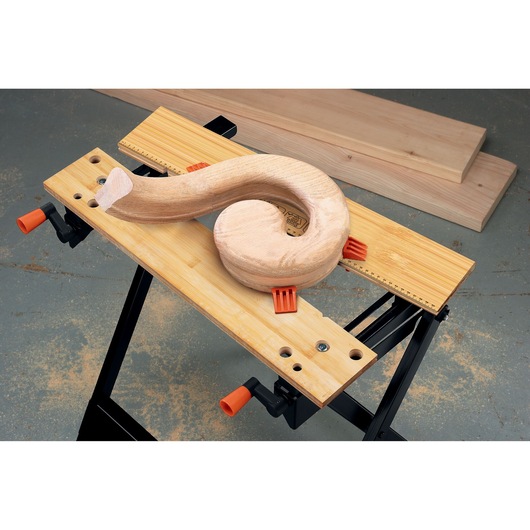 A BLACK+DECKER WM301 workbench holds a curved wooden piece securely with orange clamps. The bench has a light wooden surface and black metal legs, with wooden boards in the background.