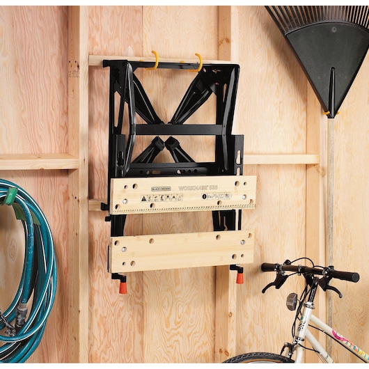 A folded BLACK+DECKER Workmate WM536 workbench is hanging on hooks attached to a wooden wall in a garage or shed. Nearby, a green garden hose, a rake, and part of a bicycle are also visible.