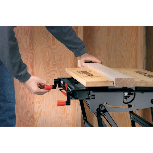 A person adjusts a BLACK+DECKER workbench, clamping a piece of wood on its surface. The workbench features black metal legs and orange handles, and is set against a wooden wall background.