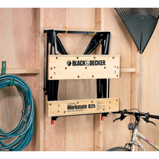 A BLACK+DECKER Workmate 825 portable workbench is folded and hung on a wall in a wooden shed. Nearby, there is a garden rake, a coiled hose, and the handlebar of a bicycle visible.