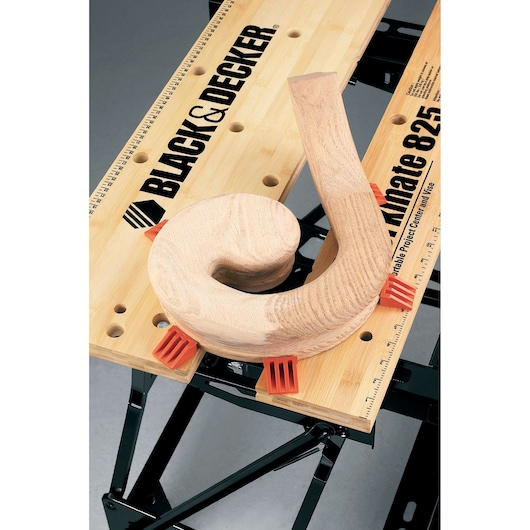 A BLACK+DECKER Workmate 825 portable workbench with a piece of curled wood clamped on the surface using orange plastic holders.